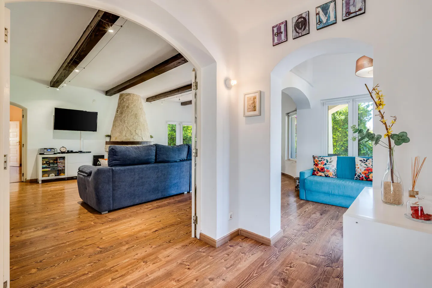 A bright, open-concept living space with hardwood floors, white walls, and arched doorways. A blue sofa and stone fireplace are visible in the living room.