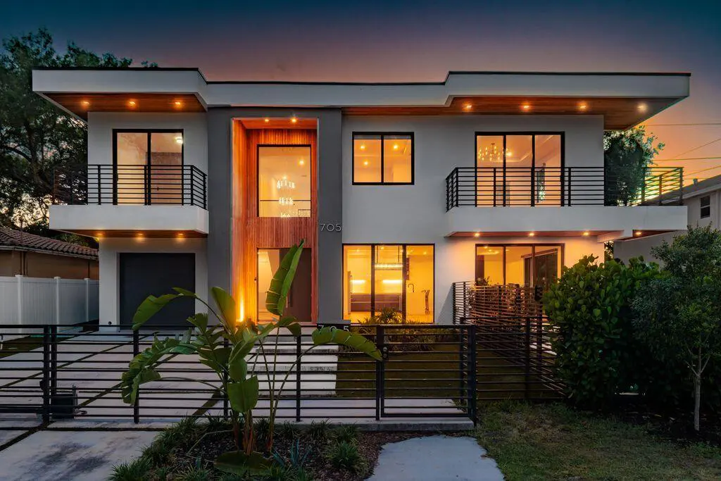 Two-story modern home with white and gray exterior, wood accents, black railings, and a black horizontal slat fence.