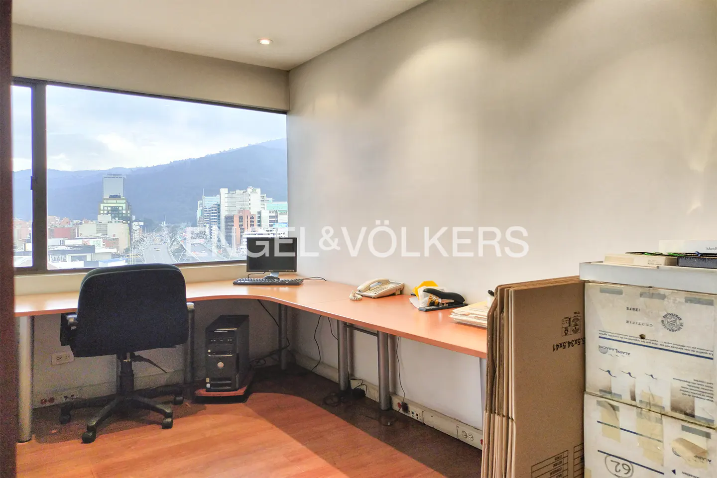 Office space with a large window view of a city and mountains. A desk with a computer, phone, and office supplies is visible.