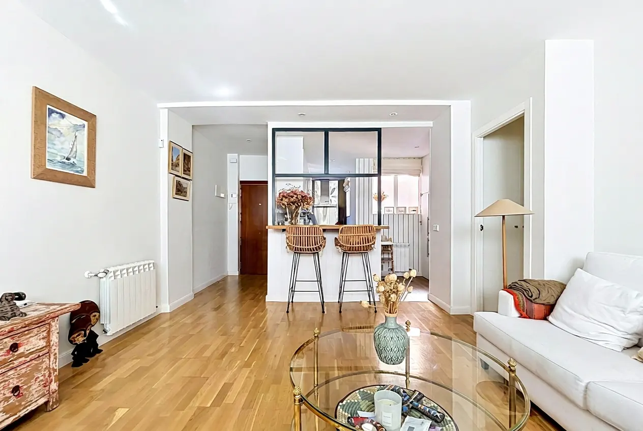 Bright living room with wood floors, white walls, and a glass coffee table. A kitchen bar with two stools is visible in the background.