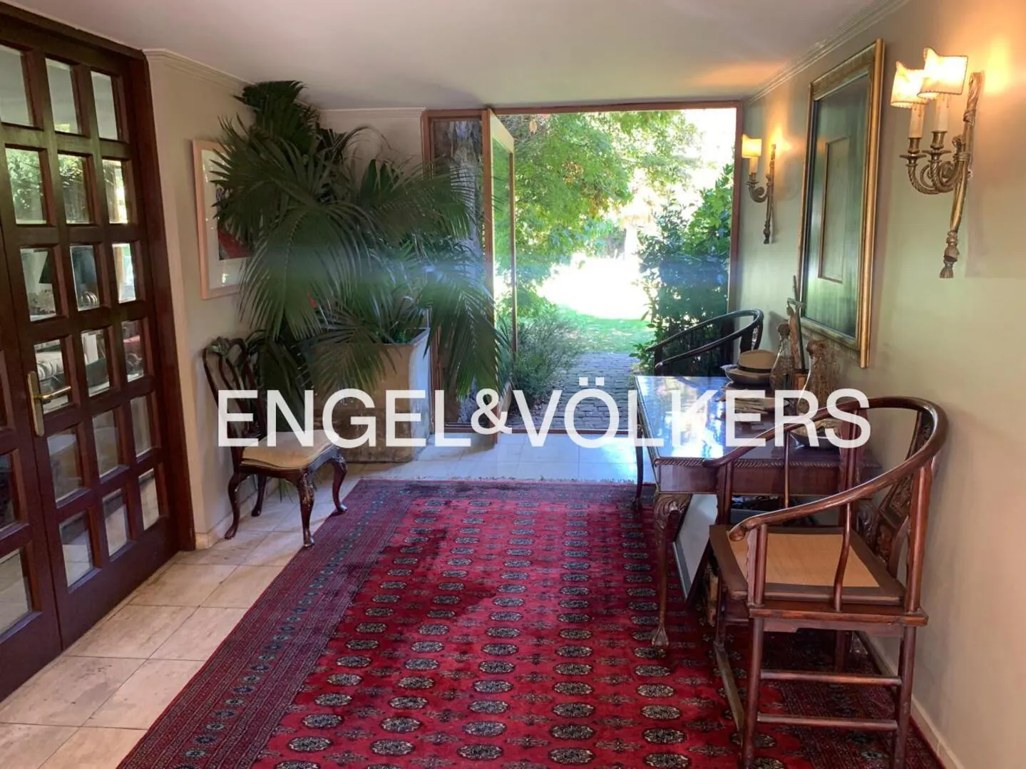 A home's entryway with a red rug, wooden chairs, and a view of a green garden through open doors.