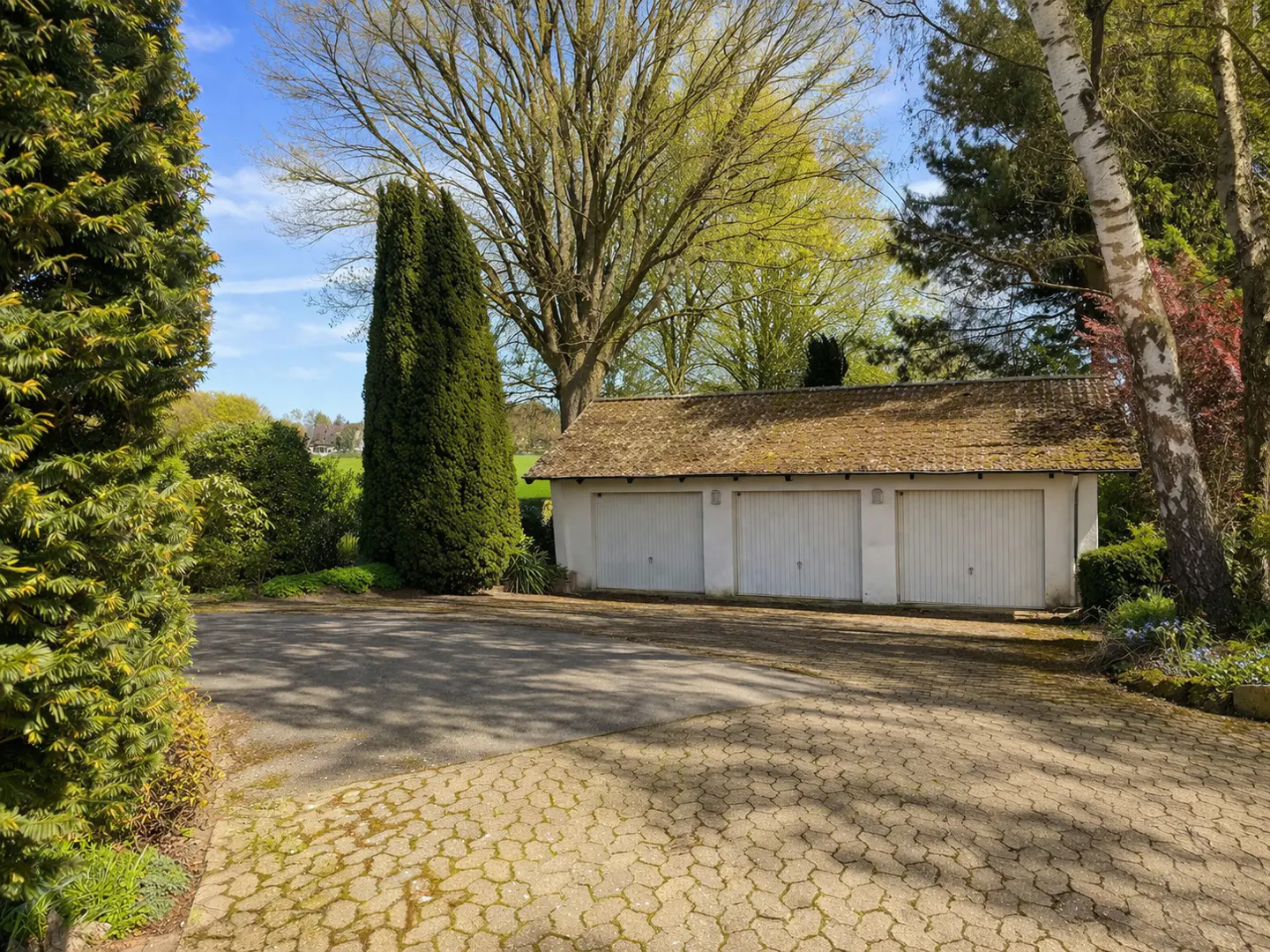 Three-car garage with white doors and a brown roof, surrounded by green trees and a paved driveway.
