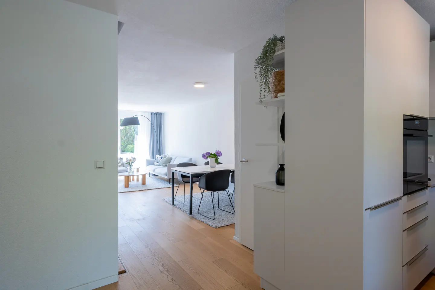 View from a white kitchen into a bright, open-plan living and dining area with light wood floors and modern furniture.