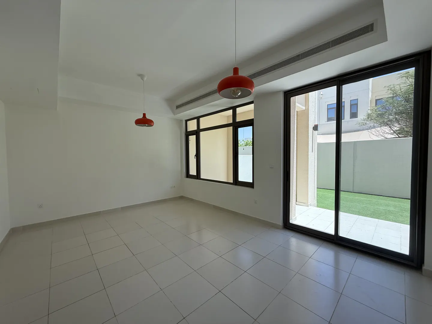 Bright, empty room with white walls, tile floor, and red pendant lights. A large sliding glass door leads to a patio and green lawn.