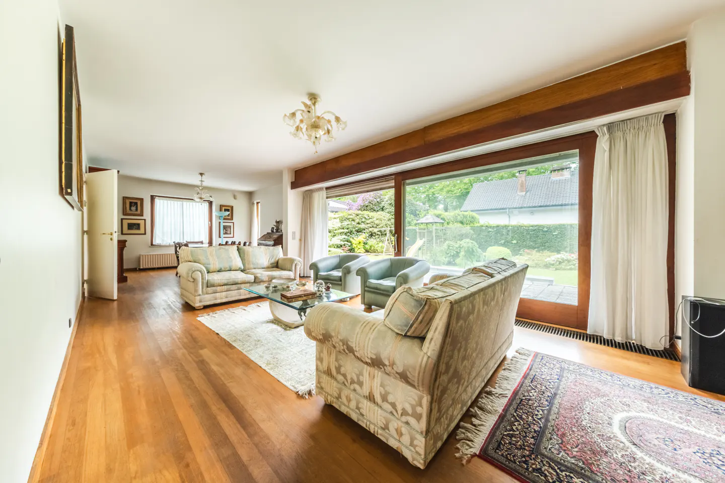 Bright living room with hardwood floors, patterned sofas, armchairs, and a large window overlooking a green garden.