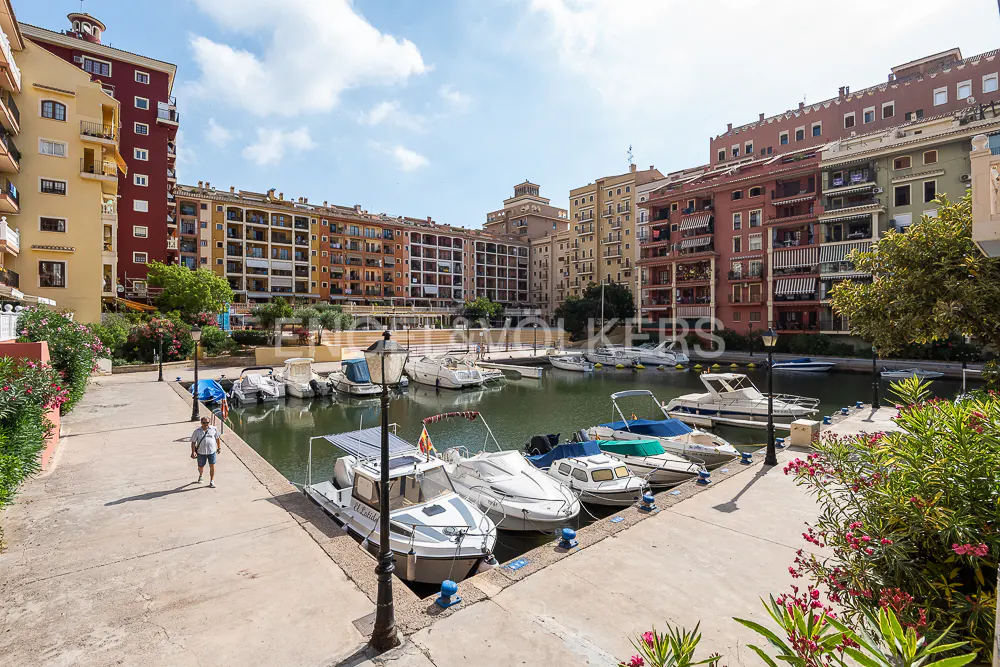 Waterfront view of a marina with boats docked along a concrete walkway, surrounded by colorful apartment buildings under a blue sky.