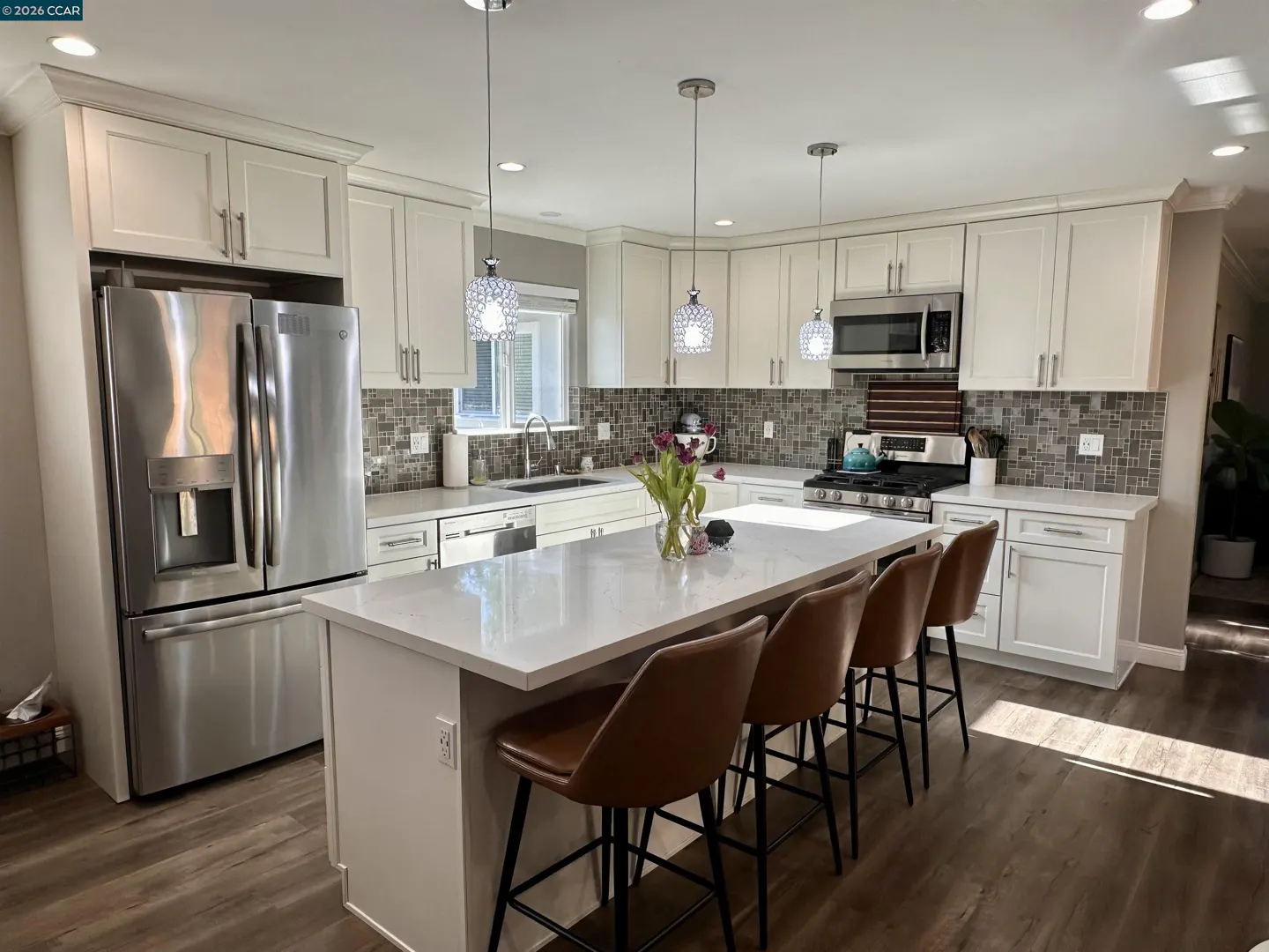 Bright kitchen with white cabinets, stainless steel appliances, and a large island with brown leather stools. Three pendant lights hang above the island.