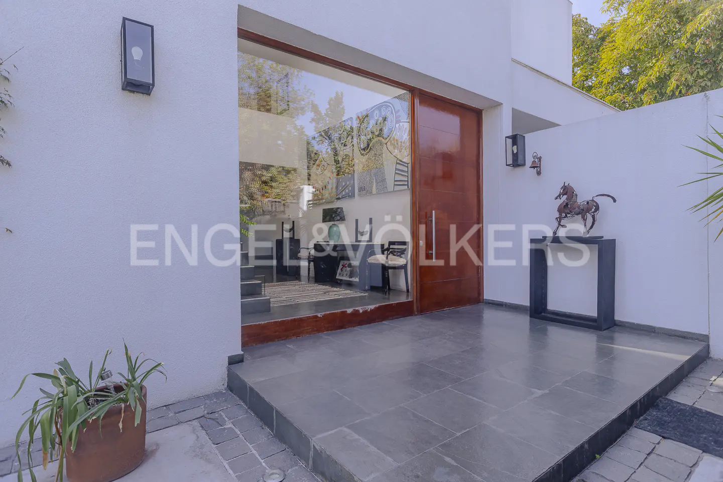 Exterior view of a modern home entrance with a large glass door and a brown wooden door. A metal horse statue sits on a black table. The Engel & Völkers logo is visible.