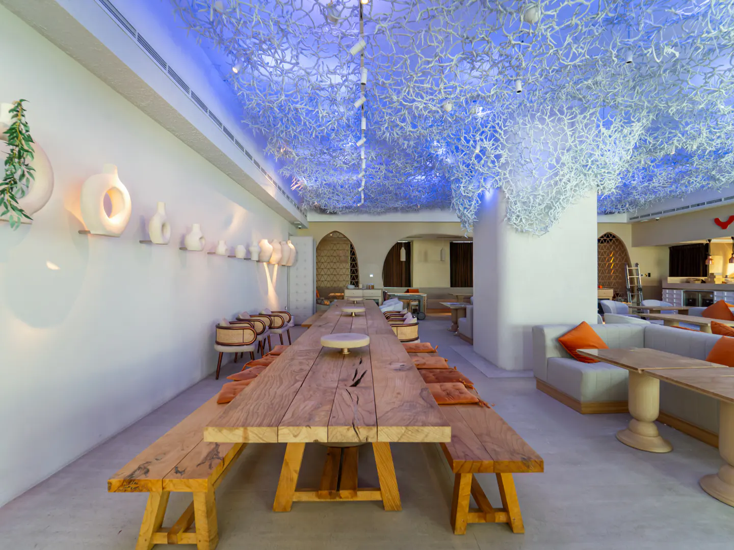 Modern restaurant interior with a long wooden table, benches, and a ceiling installation lit by blue ambient lighting.