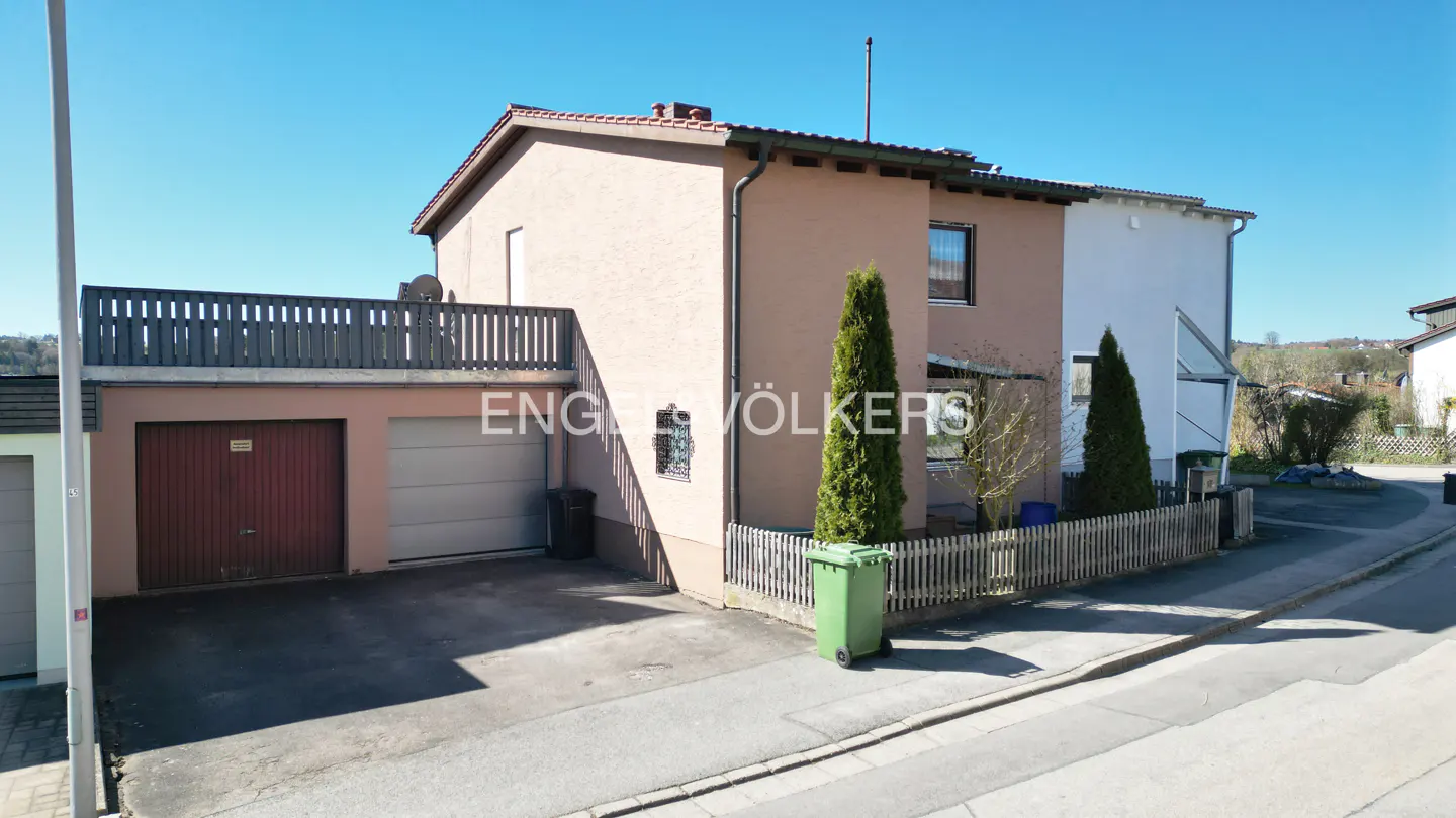 Exterior view of a two-story house with a garage, brown stucco, and a small yard with green bins.