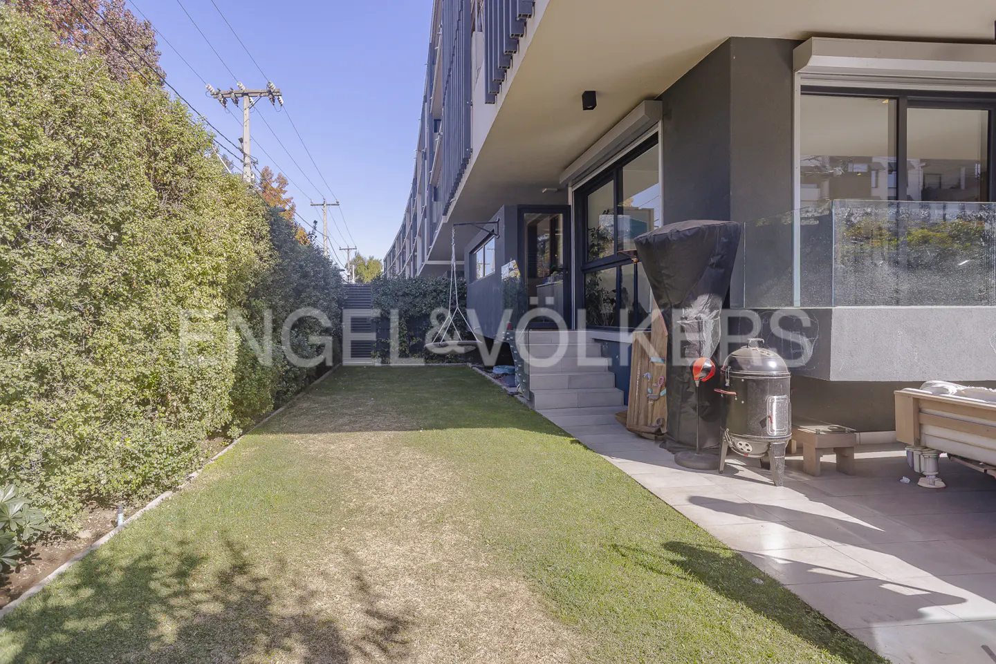 Exterior view of a modern, multi-story home with a green lawn, patio, and grill. Tall green hedges line the property.