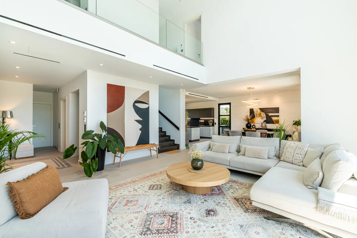 Bright, modern living room with white walls, a large abstract painting, and a light gray sectional sofa. A round wooden coffee table sits on a patterned rug.