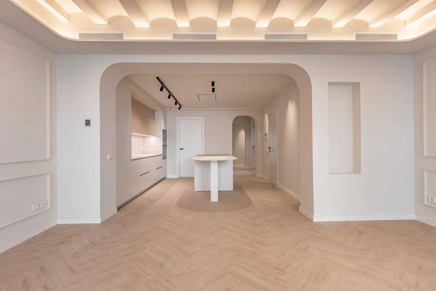 Bright, modern apartment interior with herringbone wood floors. White walls, arched doorways, and a kitchen with a center island are visible.