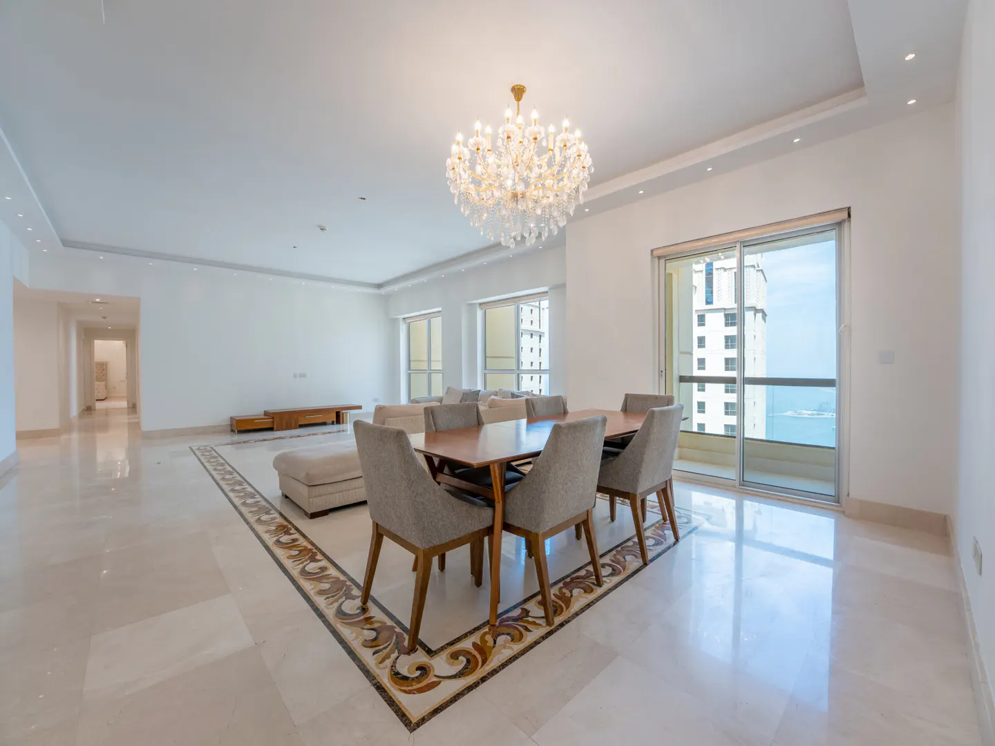 Bright, open-plan living and dining area with marble floors, a wooden table with six chairs, and a crystal chandelier. Balcony with ocean view.