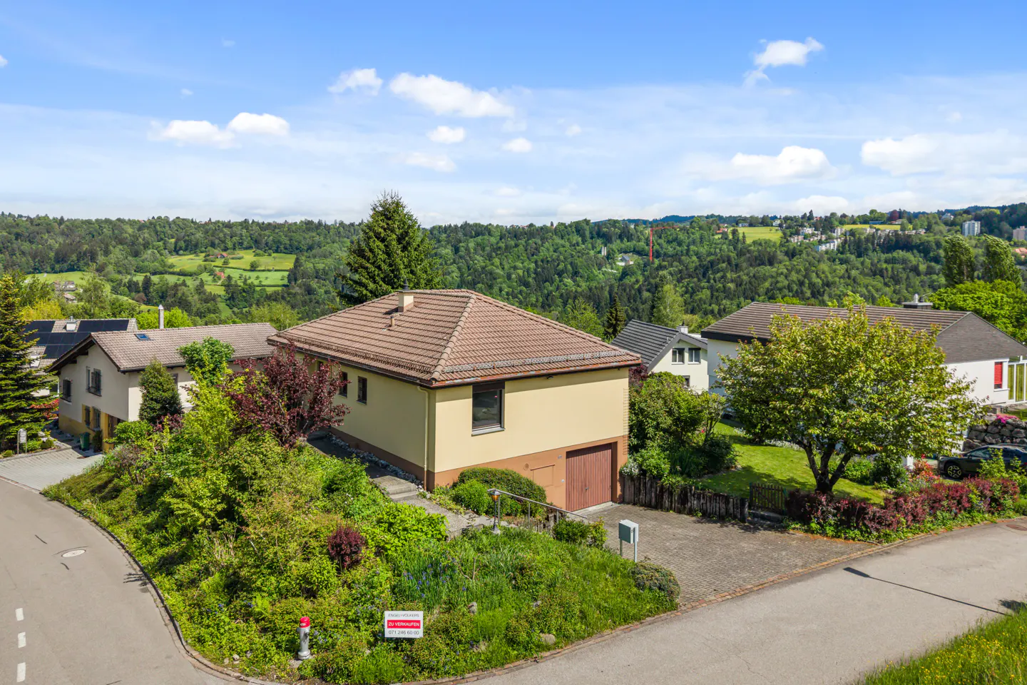 Tan house with brown trim and roof, garage, and driveway. Lush green trees and hills in the background under a blue sky.
