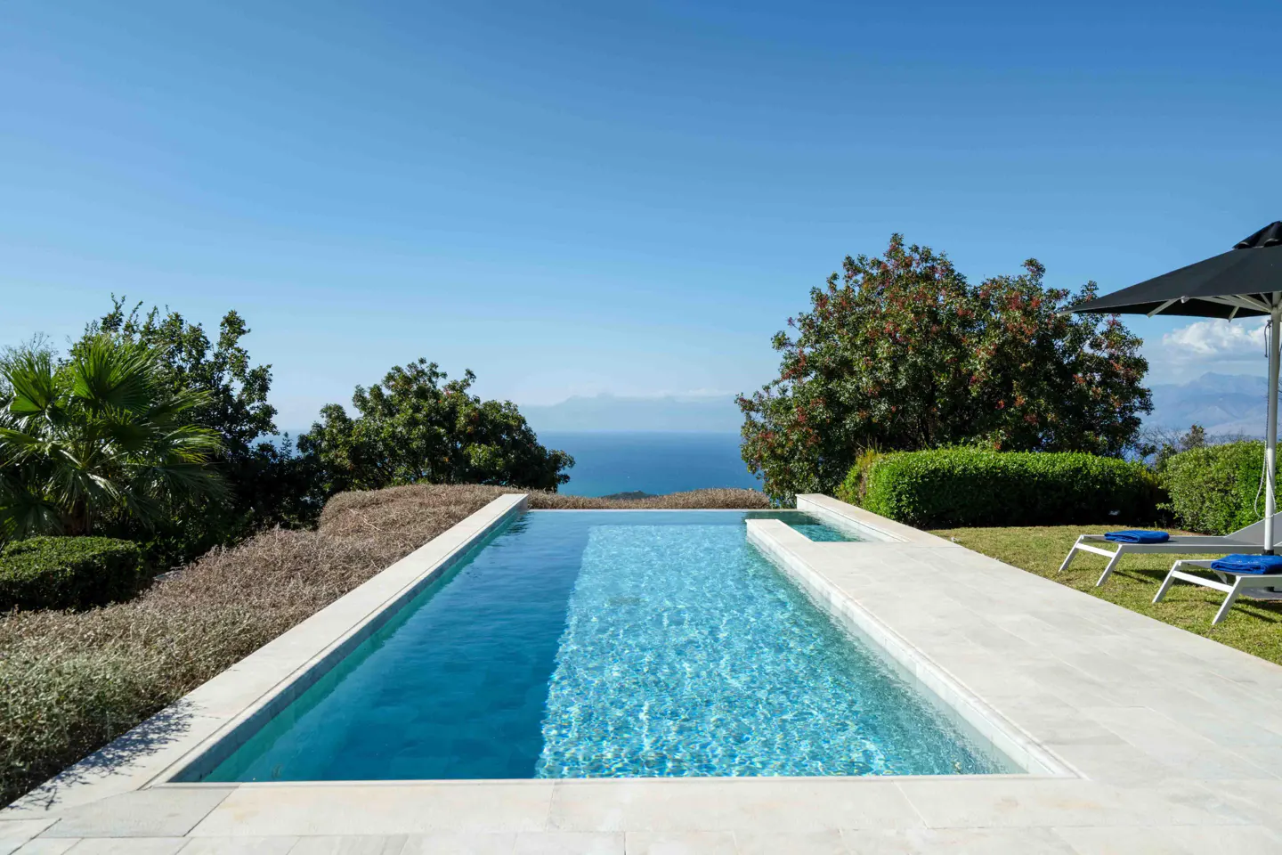 A rectangular pool with clear blue water overlooks the ocean under a clear blue sky.