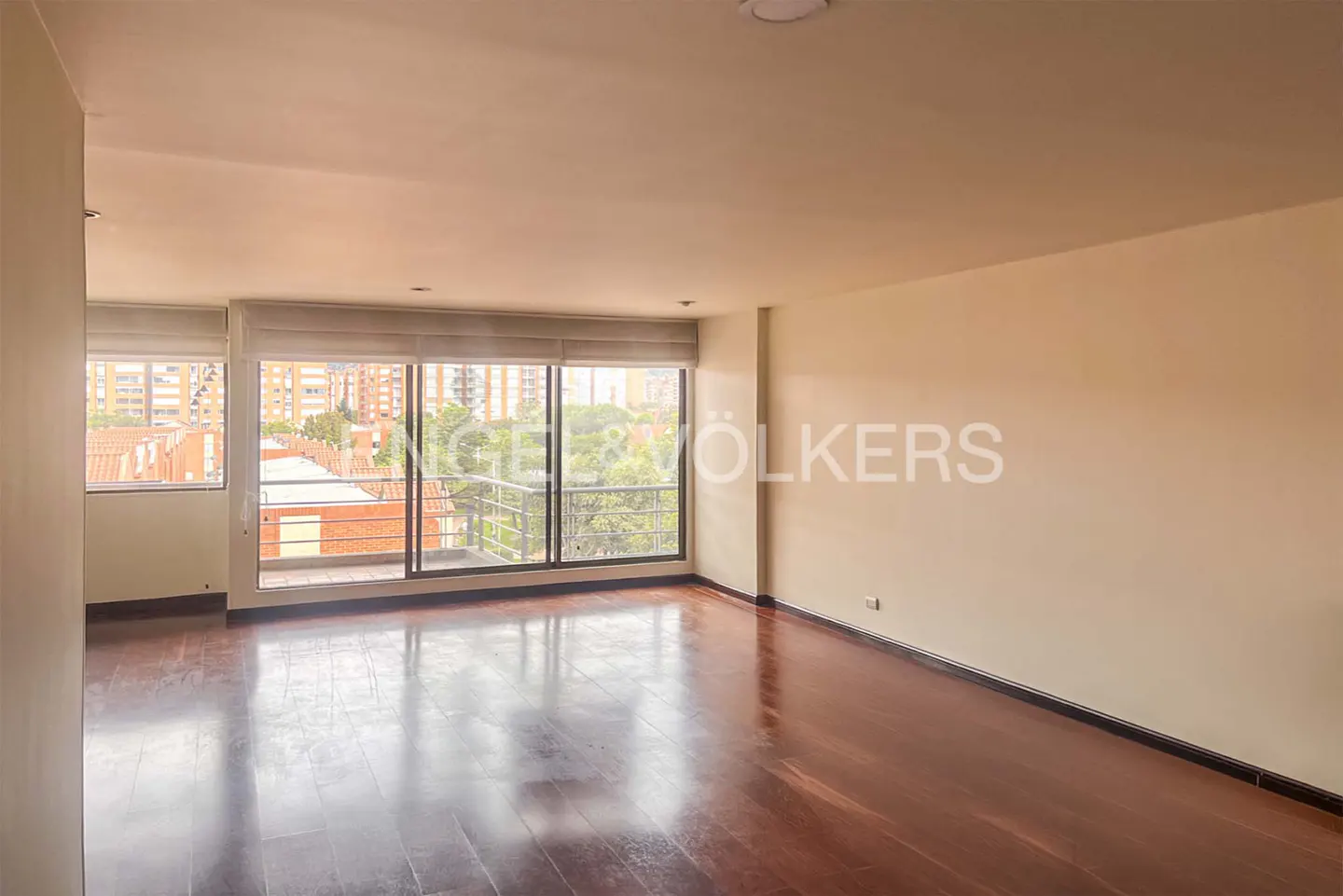 Empty room with hardwood floors and large windows showing city views. Cream-colored walls and natural light fill the space.