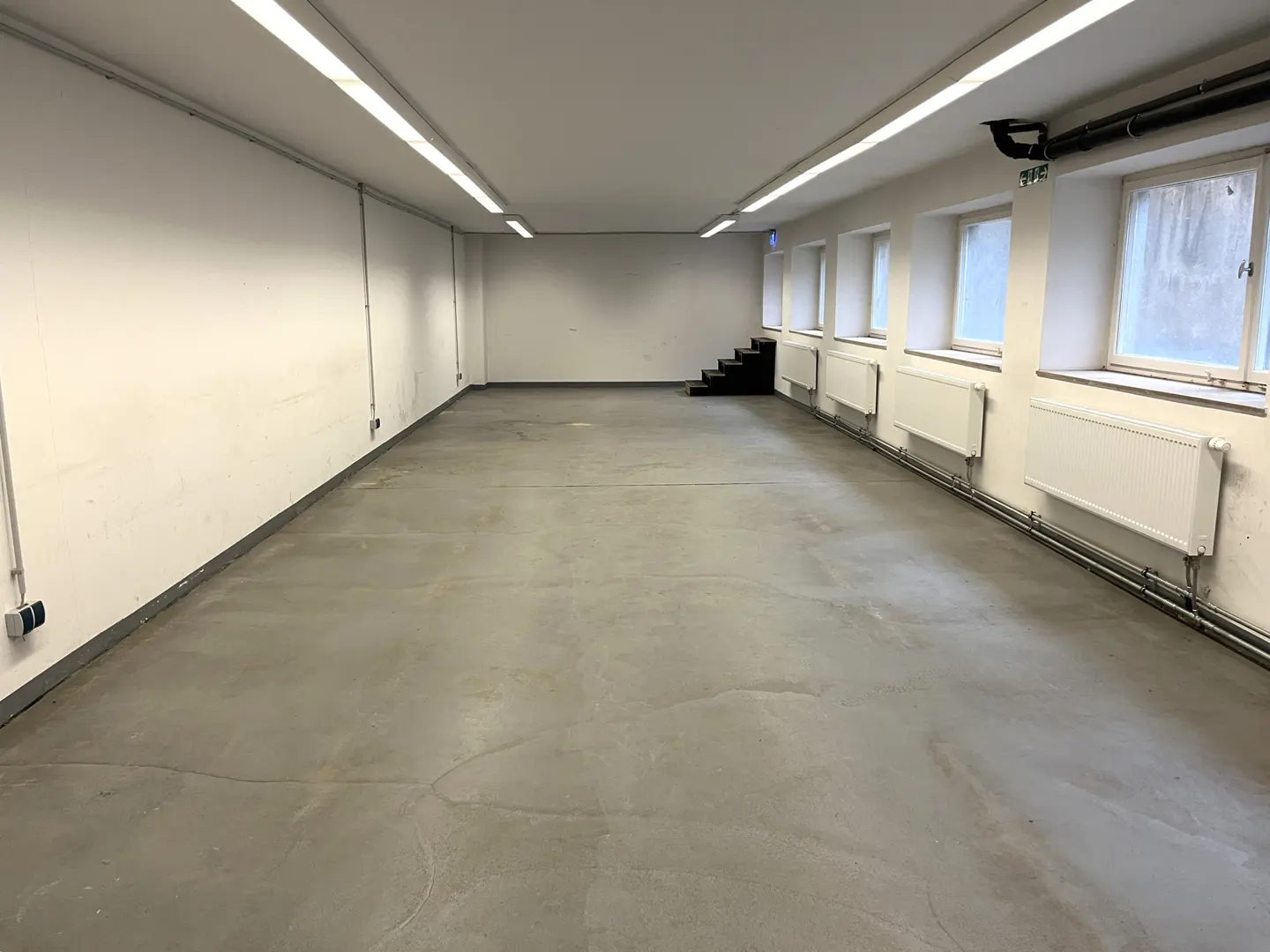 Empty room with gray floor, white walls, and fluorescent lighting. Windows line the right wall above white radiators. Black stairs in the back.