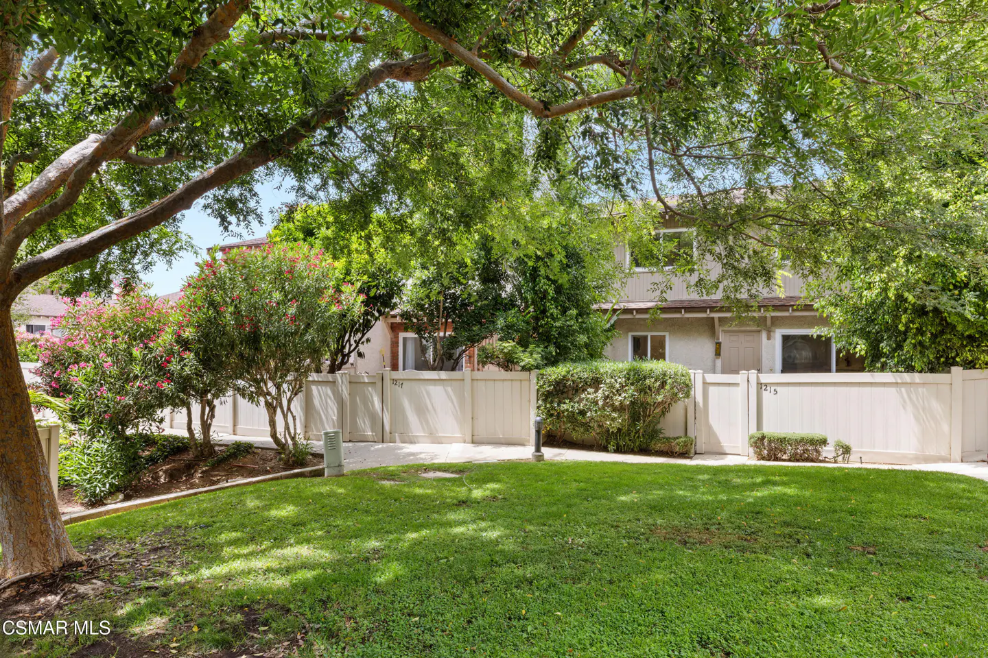 Exterior view of a two-story townhouse with a white fence, green lawn, trees, and pink flowers.