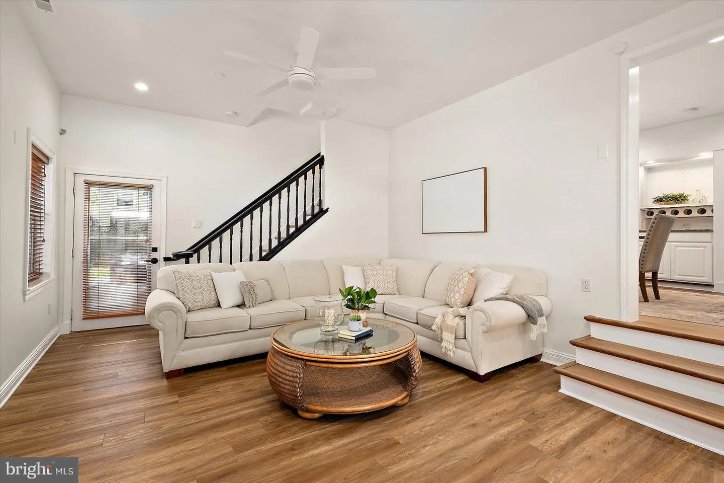 Bright living room with wood floors, white walls, and a beige sectional sofa. A round wicker coffee table sits in front of the sofa. Stairs lead to a kitchen.