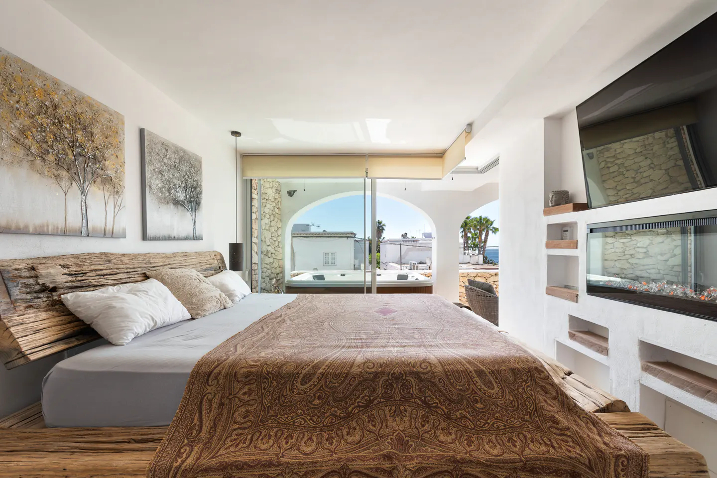 Bedroom with a rustic wood bed frame, paisley blanket, and a view of a jacuzzi and ocean. Modern fireplace on the right.
