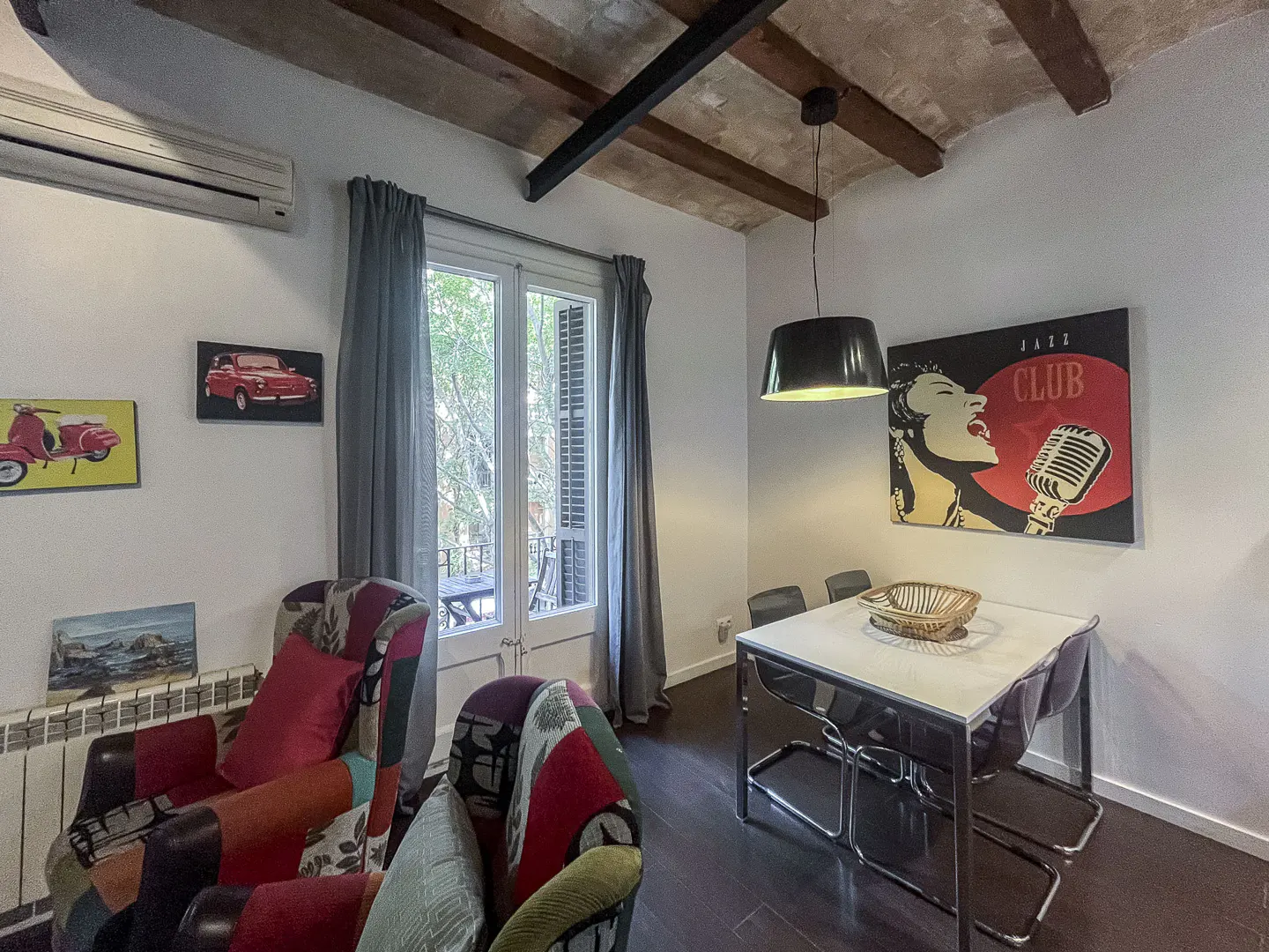 Dining area with a white table, modern chairs, a jazz club wall art, and colorful patterned armchairs under wood beams.