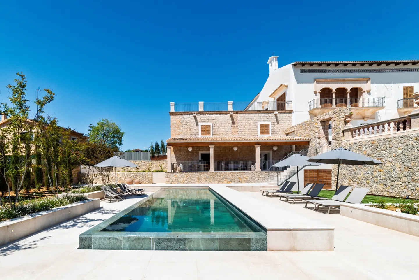 Luxury home exterior with a rectangular pool, lounge chairs, and umbrellas under a clear blue sky. The house is stone and white.