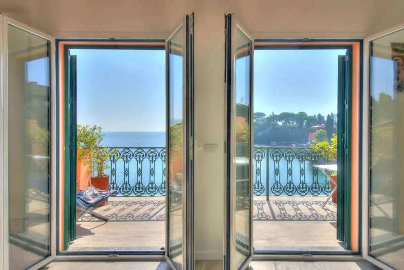 Open glass doors lead to a balcony with a blue ocean view. The balcony has a decorative railing, plants, and furniture.
