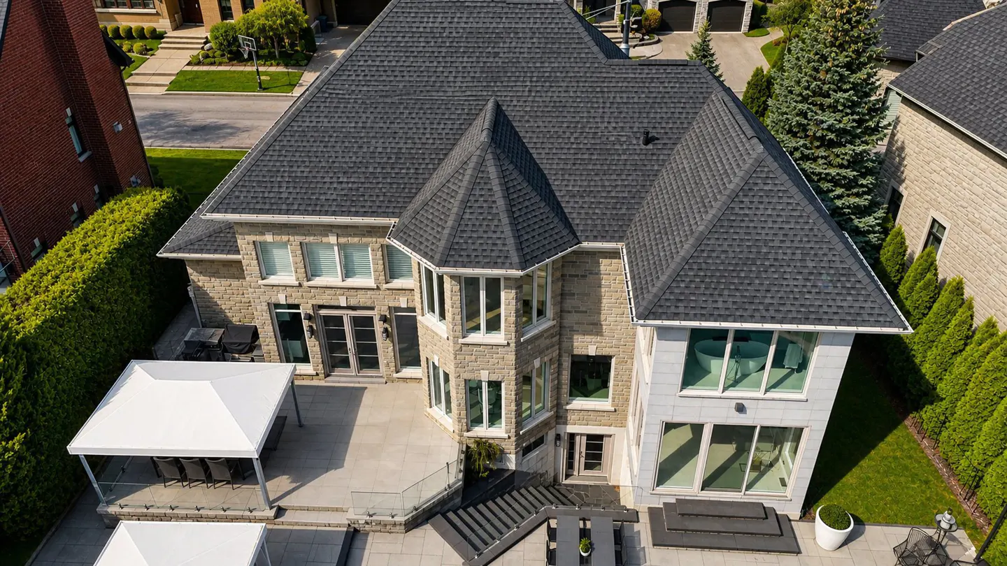 Aerial view of a large, two-story stone house with a dark gray roof, a patio with white canopies, and green landscaping.