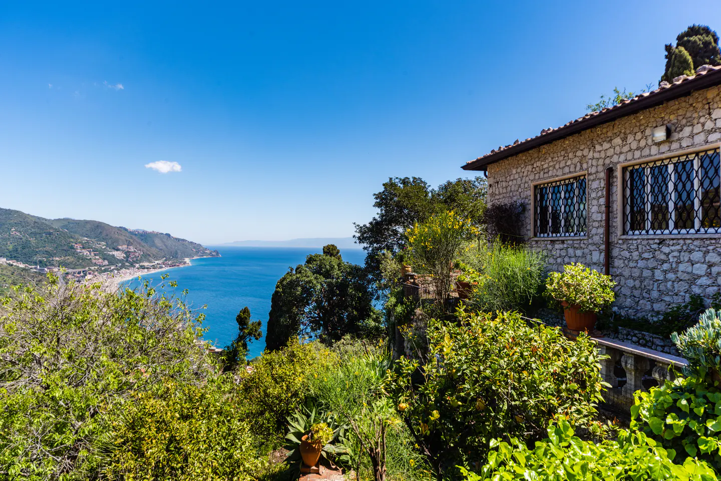 Stone house with barred windows overlooks the blue sea and green hills under a clear blue sky. Lush greenery surrounds the house.