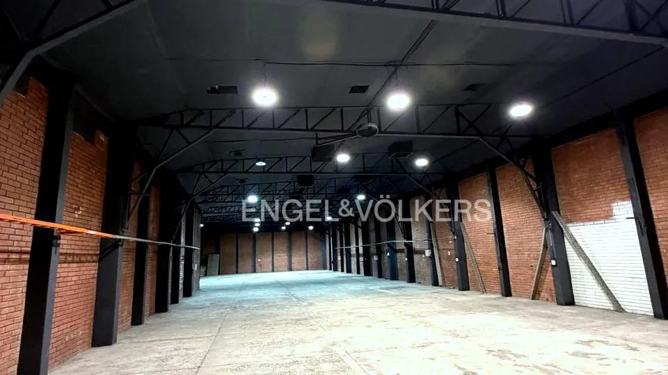 An empty warehouse with brick walls, black steel beams, and a concrete floor. Overhead lighting illuminates the space.