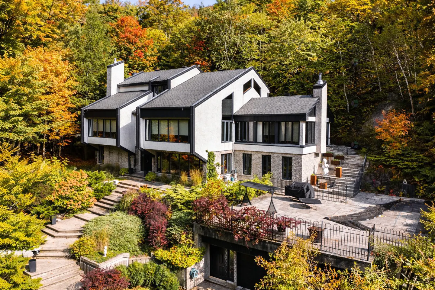 Modern white house with black trim, surrounded by colorful fall foliage. Stone steps lead up to the house and a patio with furniture.