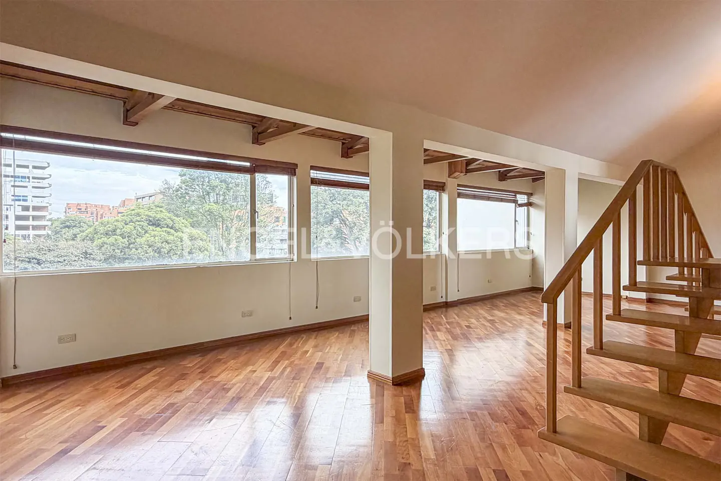 Bright, empty room with hardwood floors, large windows, and a wooden staircase. Outside the windows are trees and buildings.