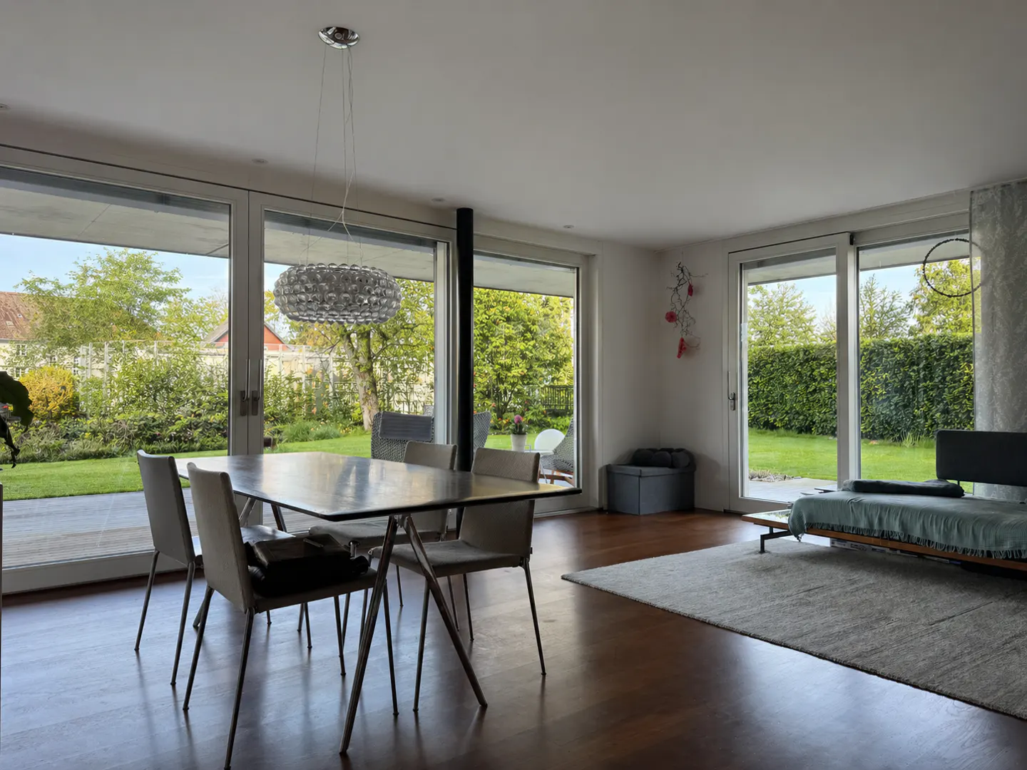 Bright, modern dining room with a table, chairs, and a decorative light fixture. Large windows overlook a green garden.