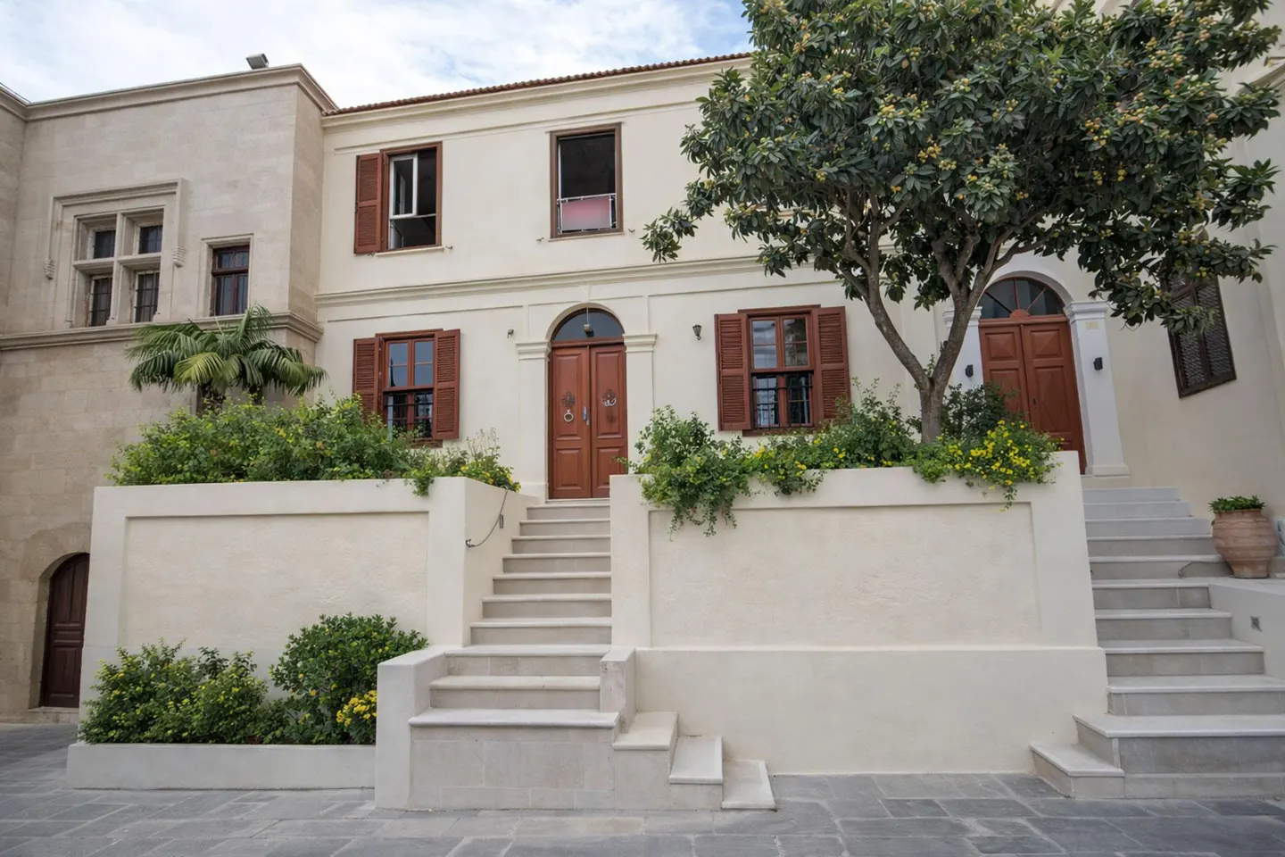 Exterior view of a two-story building with white walls, brown shutters, and a wooden door with stairs leading up to it. A tree is on the right side.