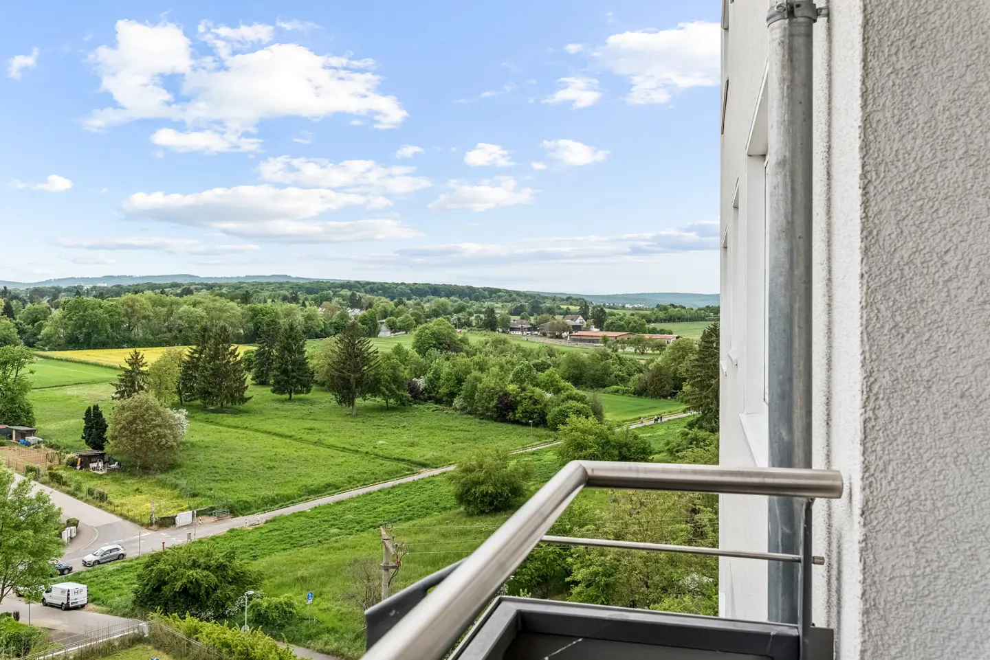 View from a balcony overlooking green fields, trees, and a blue sky with white clouds.