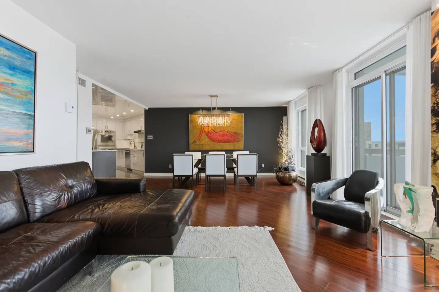 Open-concept living and dining area with dark brown leather sofa, wood floors, and large windows. Dining table with white chairs.