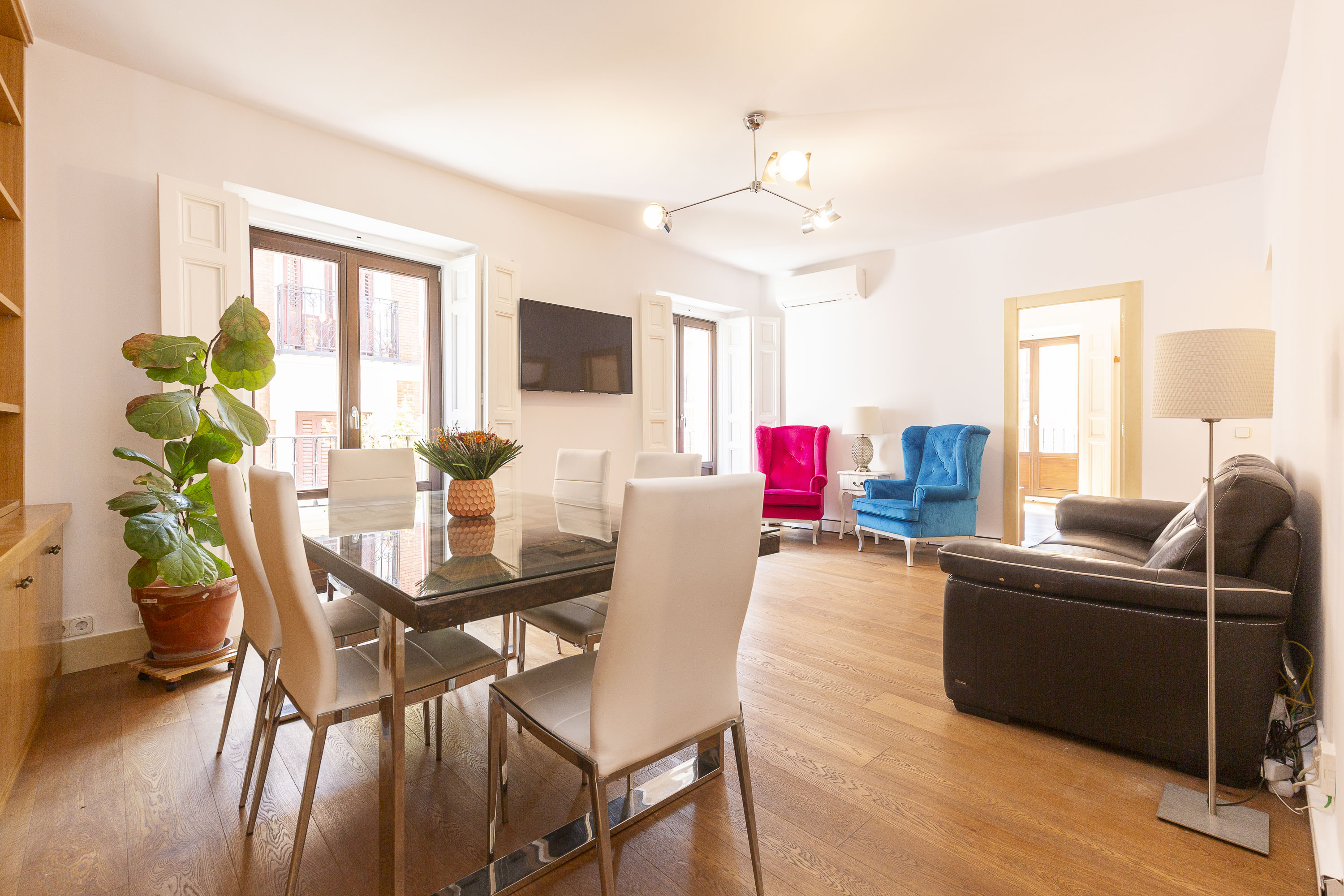 Bright living room with a glass dining table, white chairs, a black leather sofa, and vibrant pink and blue armchairs. [AI-generated text]