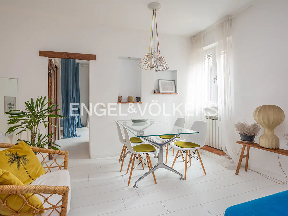Bright, airy room with white walls and floors. A glass table with four chairs sits under a colorful pendant light. A rattan sofa with yellow pillows is on the left.