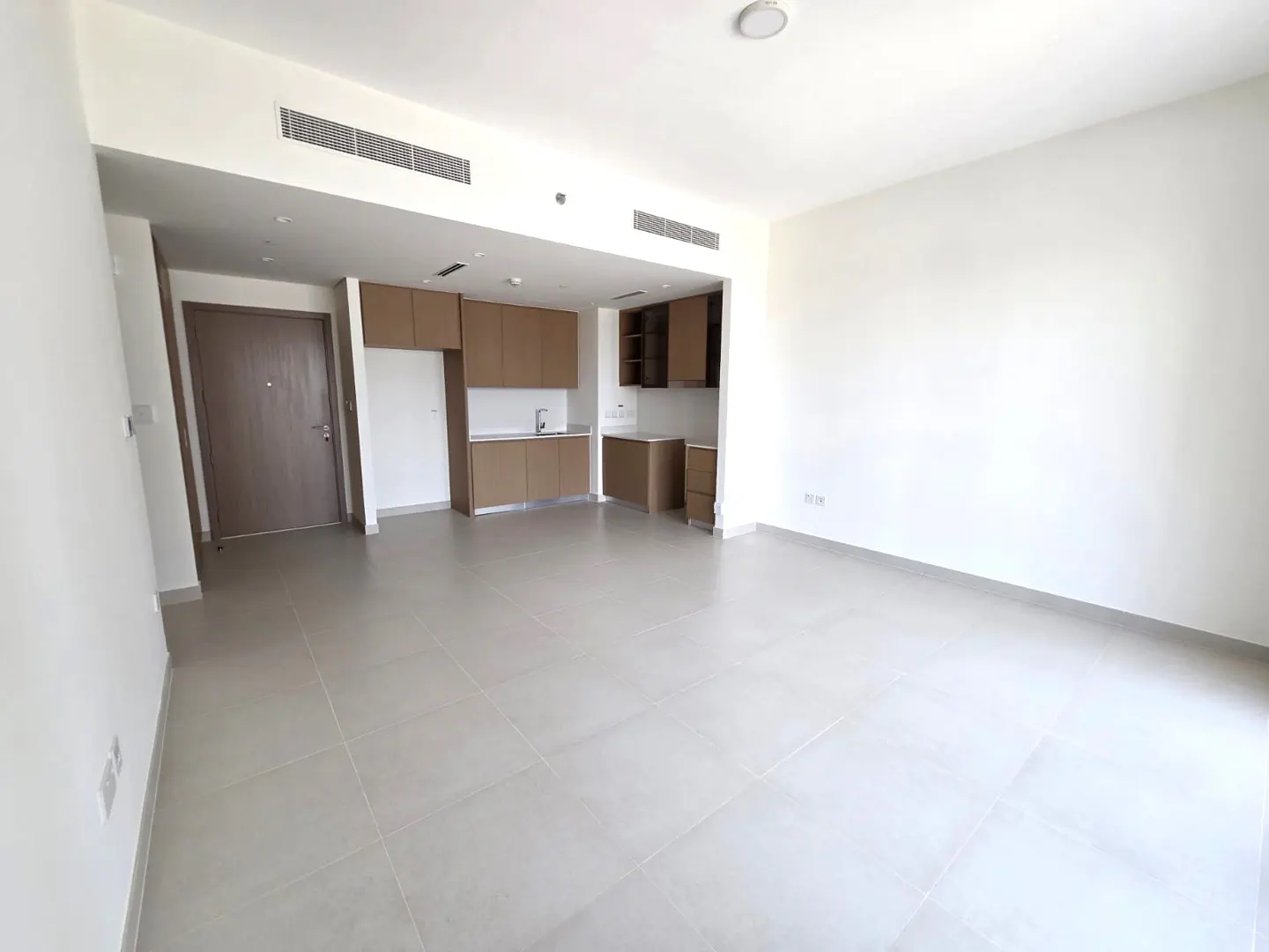 Bright, empty apartment with white walls, gray tile floor, and brown kitchen cabinets. A brown door is on the left.