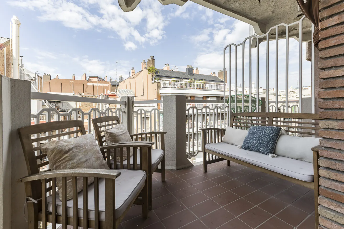 Balcony with two wooden chairs, a bench with pillows, and a view of city buildings under a blue sky.