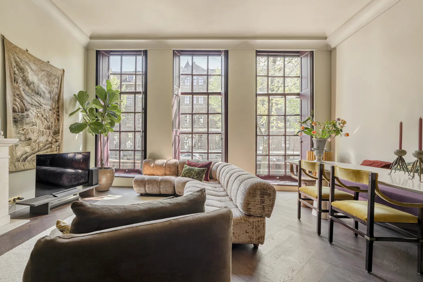 Living room with a beige sofa, brown armchair, and dining table with yellow chairs. Three large windows overlook a city street.