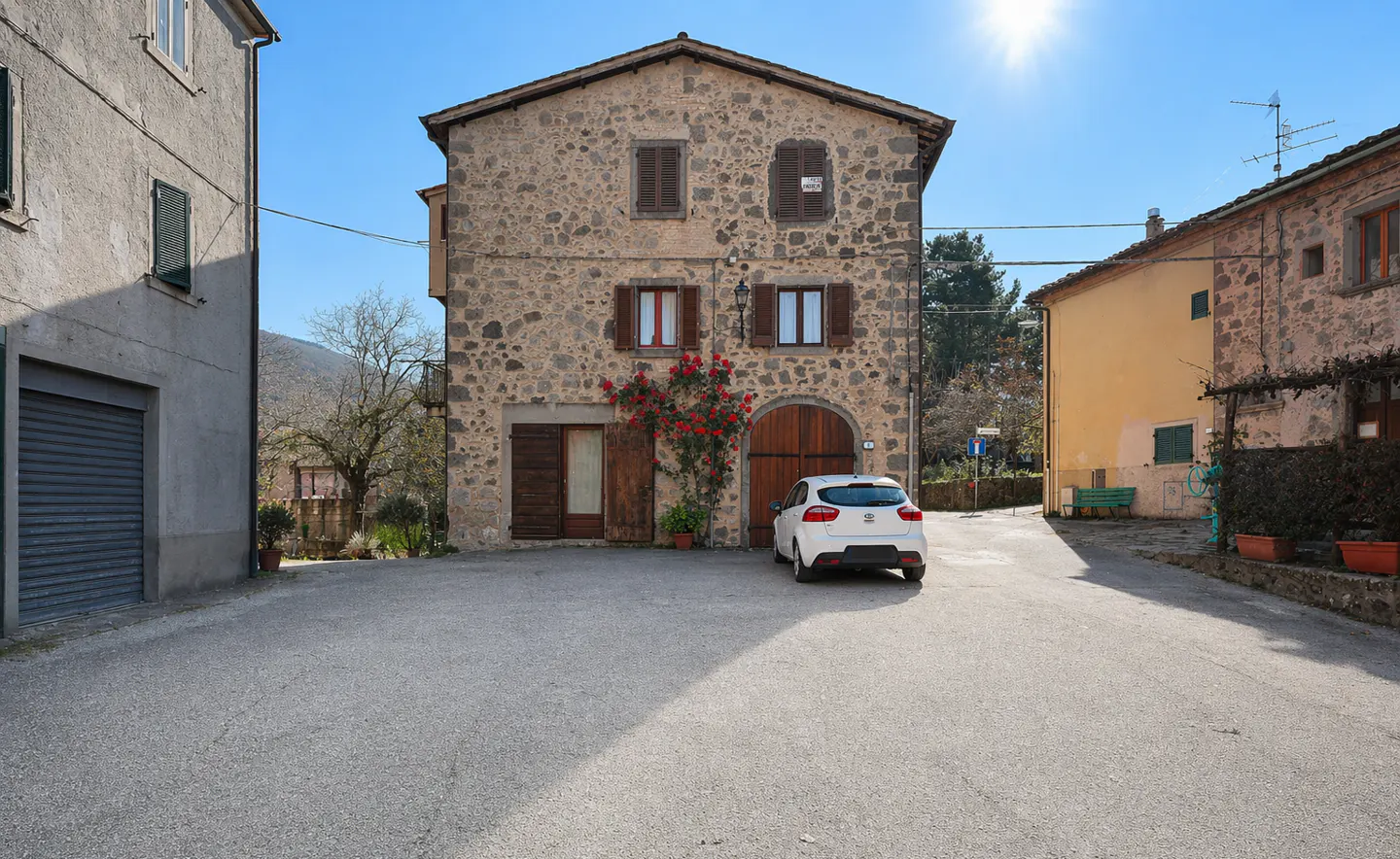 Stone house with brown shutters and a red rose bush, a white car parked in front, on a sunny day.