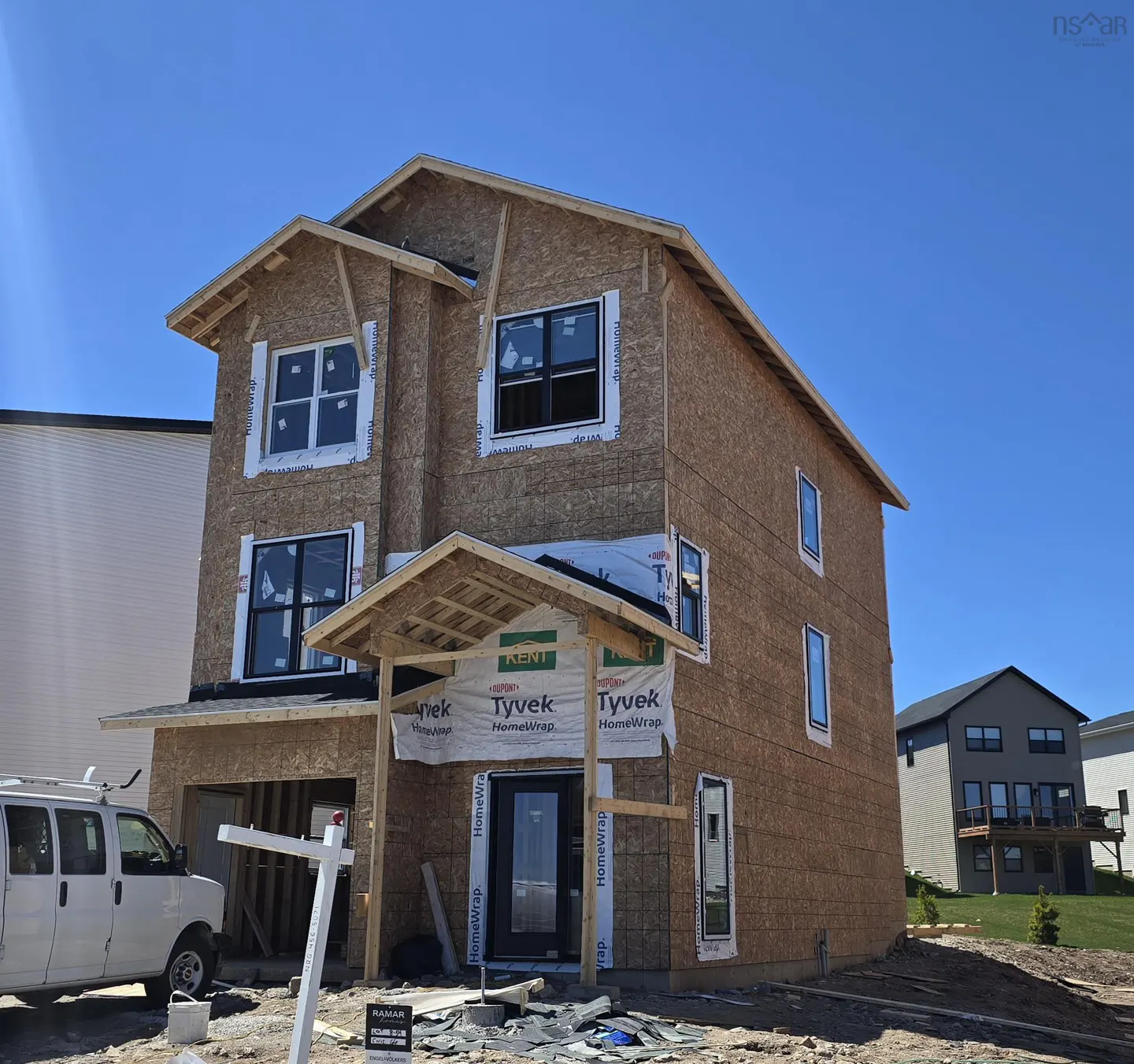 A two-story house under construction with Tyvek wrap and black windows under a clear blue sky. A white van is parked nearby.