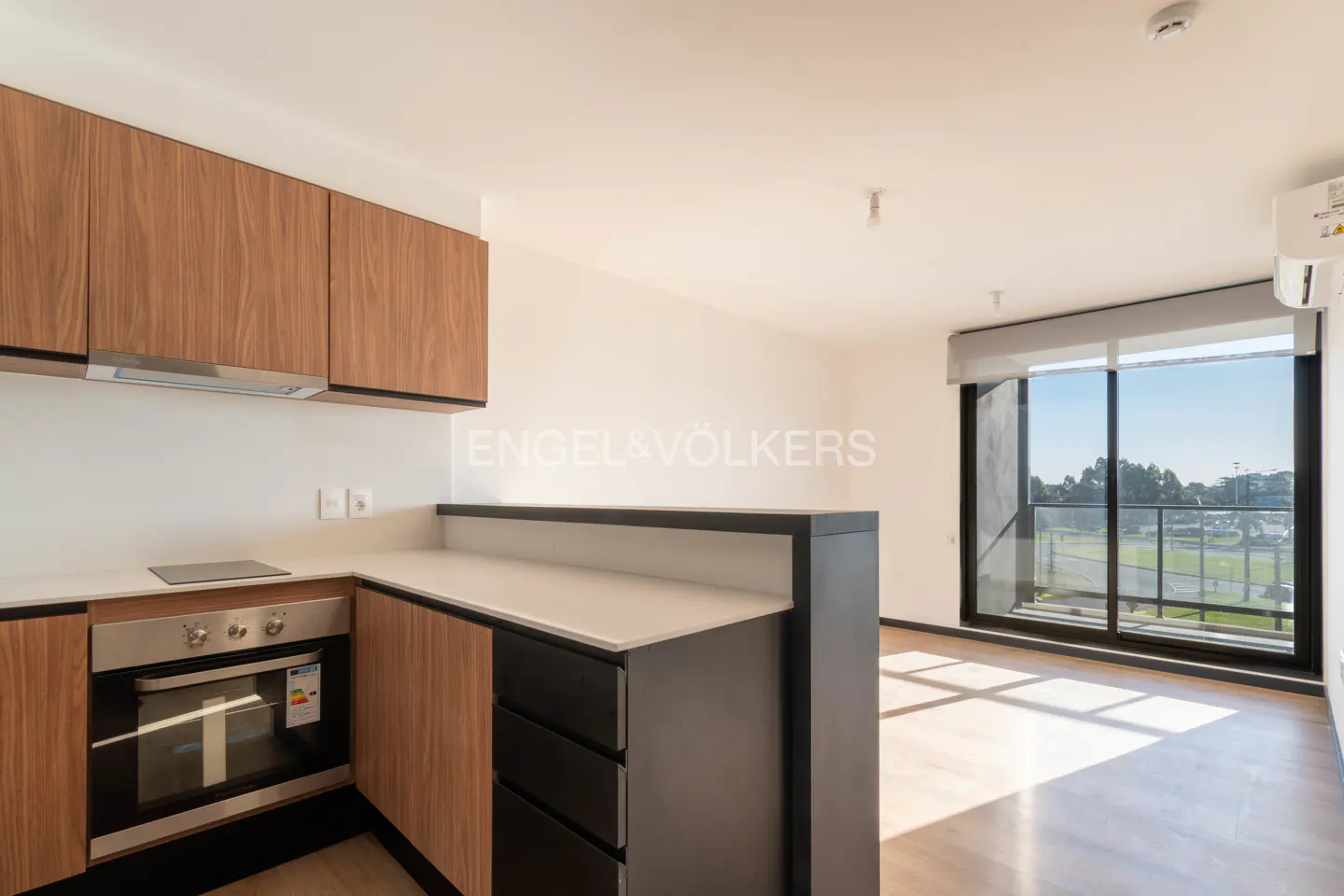 A modern kitchen with wood cabinets, a white countertop, and a stainless steel oven. A large window offers a view of the outside.