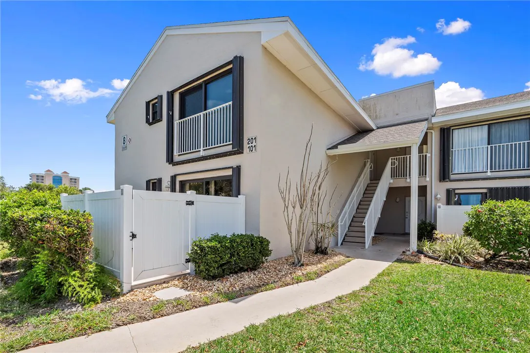 Exterior view of a two-story condo with a white fence, green lawn, and blue sky. The building is beige with white trim and black shutters. A walkway leads to the entrance.