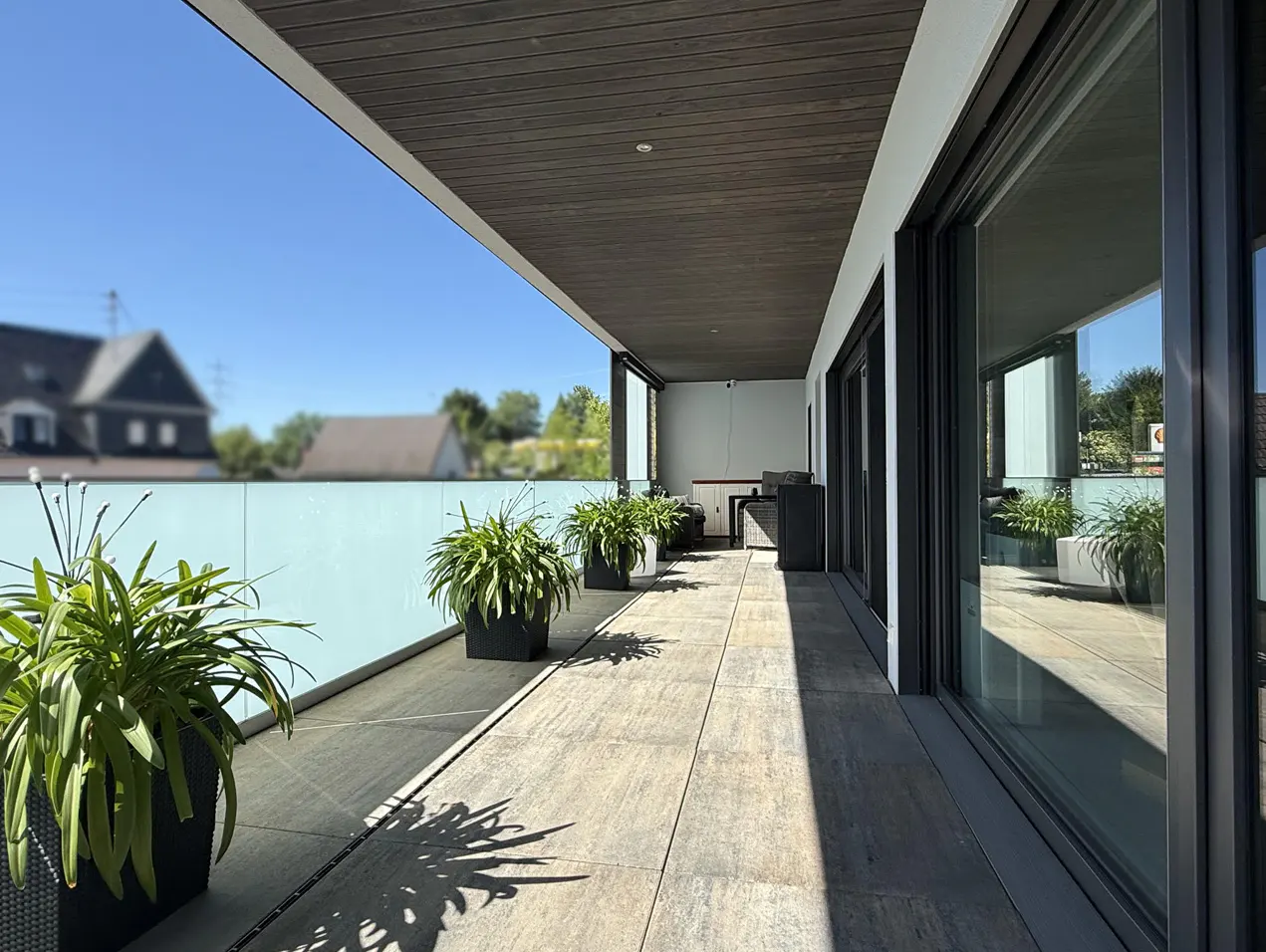 A modern balcony with potted plants, sliding glass doors, and a gray wood ceiling. A light blue glass railing offers privacy.