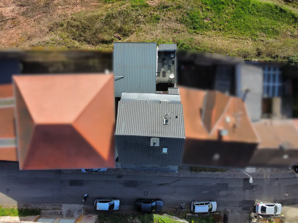 Aerial view of buildings with red and gray roofs, a road with parked cars, and a grassy hill in the background.