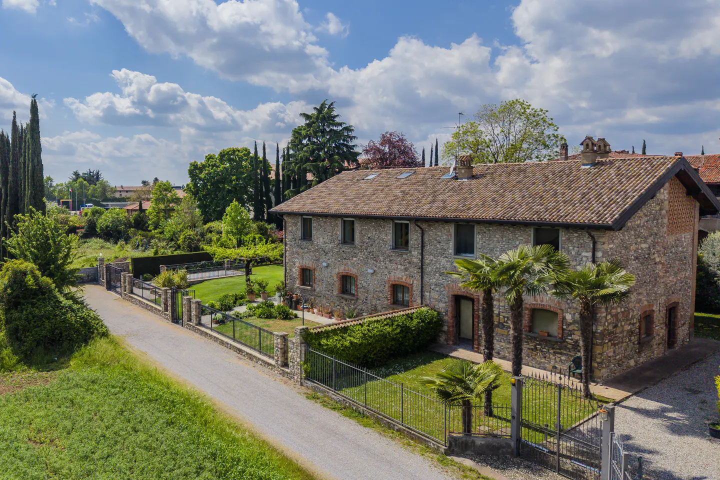 Aerial view of a two-story stone house with a red tile roof, surrounded by green trees and a lawn, under a blue sky with white clouds.