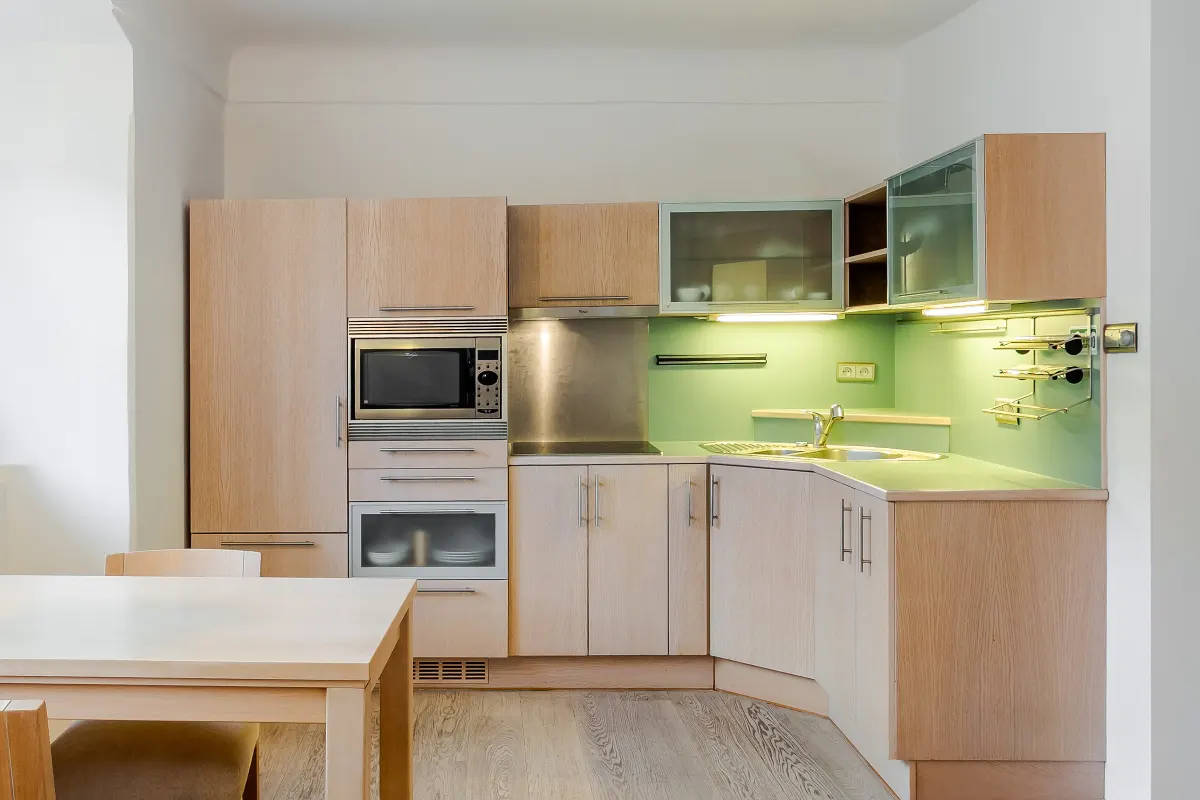 A bright kitchen with light wood cabinets, stainless steel appliances, and a green backsplash. A light wood table is in the foreground.